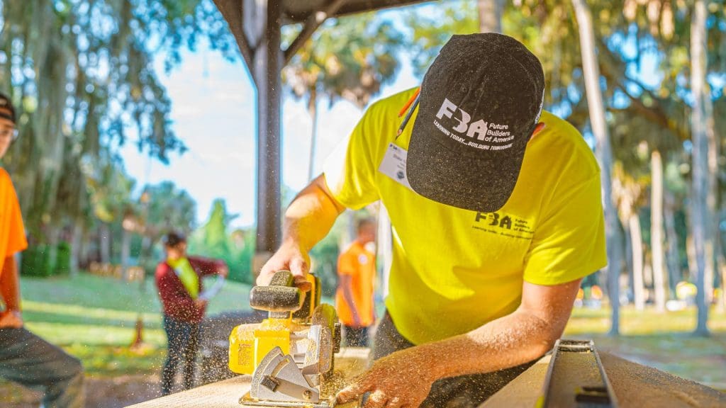 teenage man with navy bluehat with FBA logo on it sawing a piece of wood with woods in background.